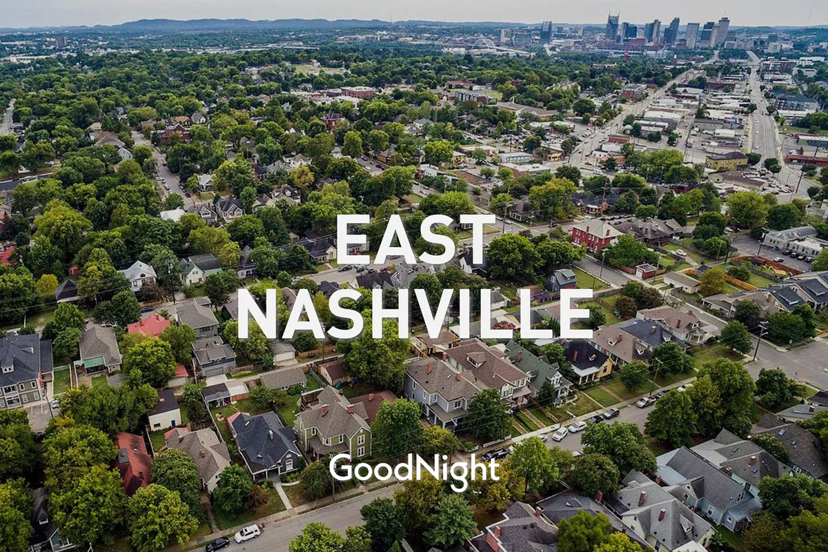 This view showcases inviting residential houses nestled among lush greenery, with the downtown Nashville skyline rising in the background. The scene captures the blend of neighborhood charm and urban excitement, with hints of sunset light enhancing the romantic mood.