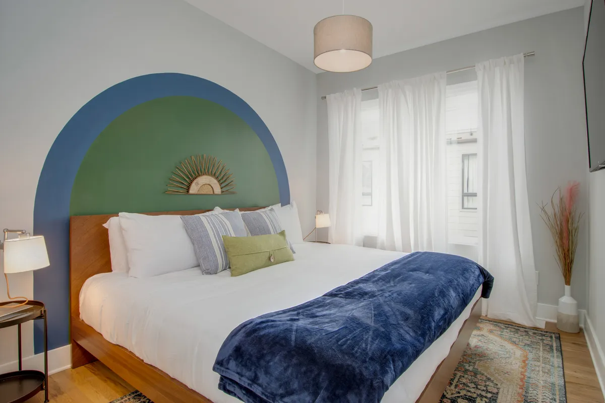 A serene bedroom featuring a bed with plush bedding, a green and blue accent wall, a decorative sunburst mirror, and light curtains softly letting in natural light. The space exudes calm with its cozy textures and stylish decor.