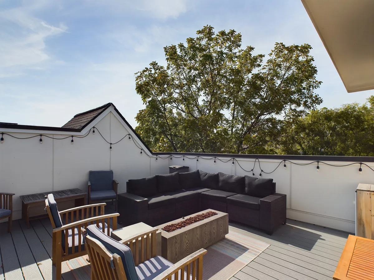 The rooftop deck is shown with a large sectional sofa and wooden chairs set within a lush space, accentuated by planter boxes and string lights. The view includes treetops and hints of the skyline, with a tranquil atmosphere perfect for lounging, reading, or enjoying coffee.