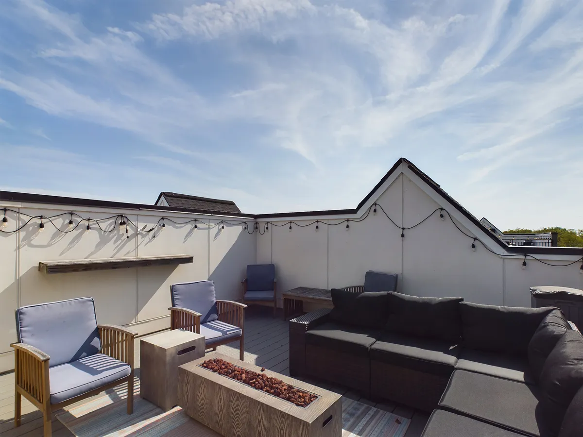 A relaxed, sociable rooftop setting is captured with an outdoor seating area positioned around a glowing fire pit. String lights crisscross overhead, enhancing the cozy, festive atmosphere. The scene suggests stargazing or hosting casual gatherings under the night sky.