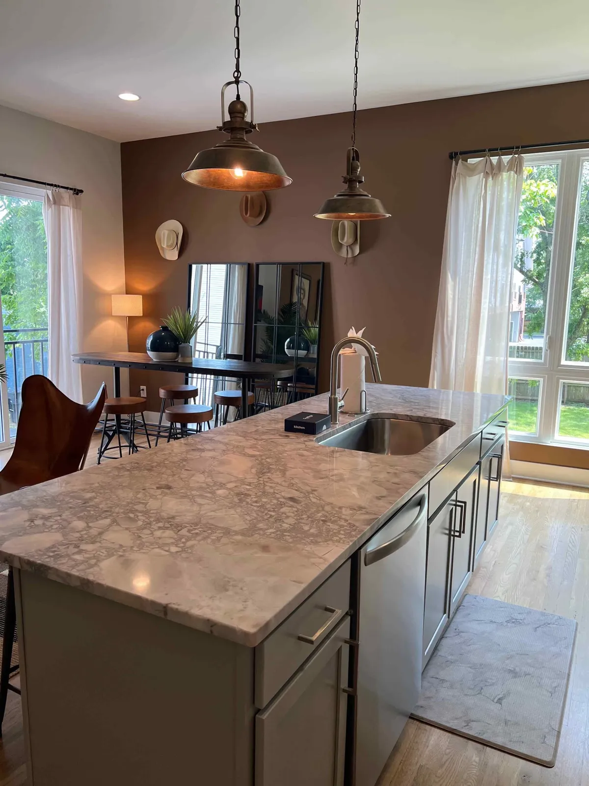 This image showcases a spacious, open-plan area featuring a modern kitchen island with sleek pendant lighting, a contemporary dining area, and sliding glass doors that bring in abundant natural light. The setting is arranged for both cooking and entertaining, with visible seating and warm, inviting tones.