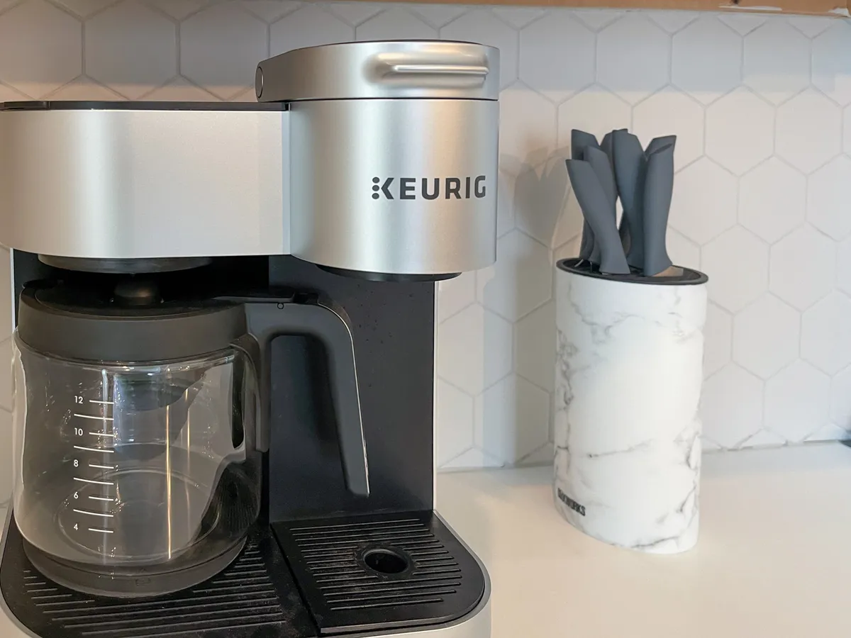 The image centers on a Keurig coffee maker and a glass coffee pot placed on a kitchen counter, with a marble utensil holder and various utensils nearby. The setup feels inviting and signals that the kitchen is ready to use upon arrival.