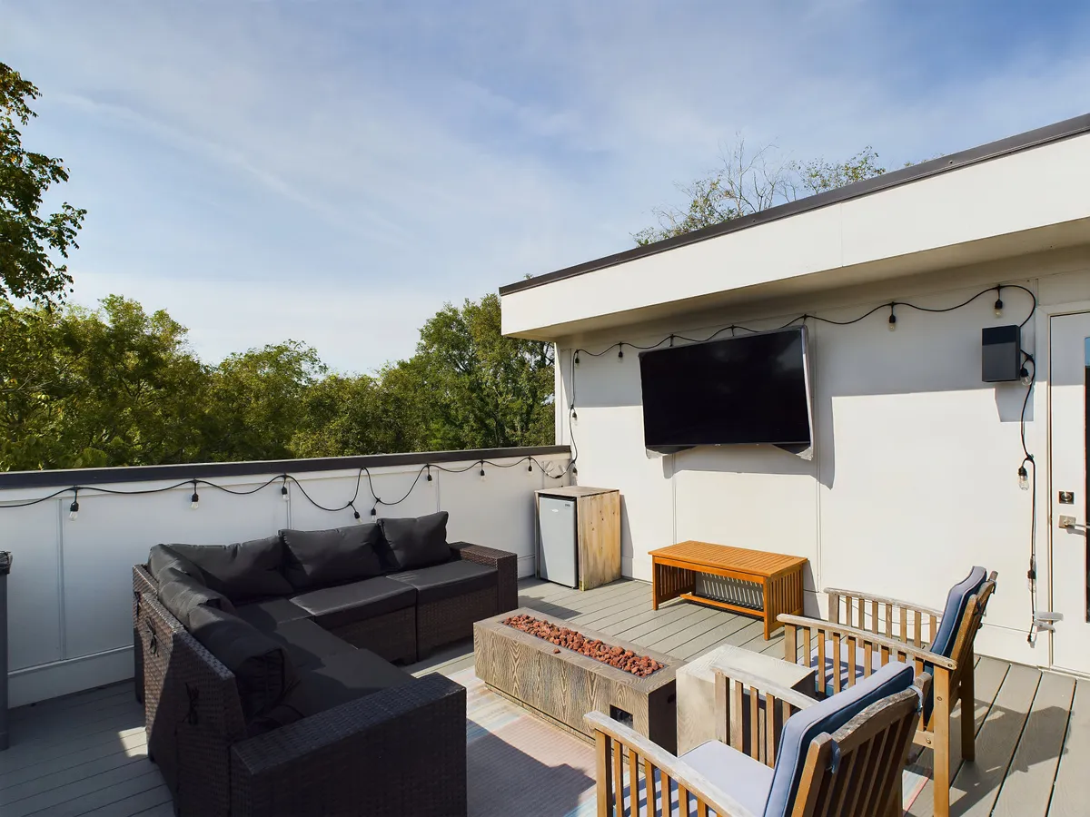 The image showcases a modern outdoor seating area surrounded by lush greenery, featuring a cozy fire pit and an outdoor television. The space is designed for relaxation and social gatherings, offering beautiful sunset views and the perfect spot for stargazing in the evening.